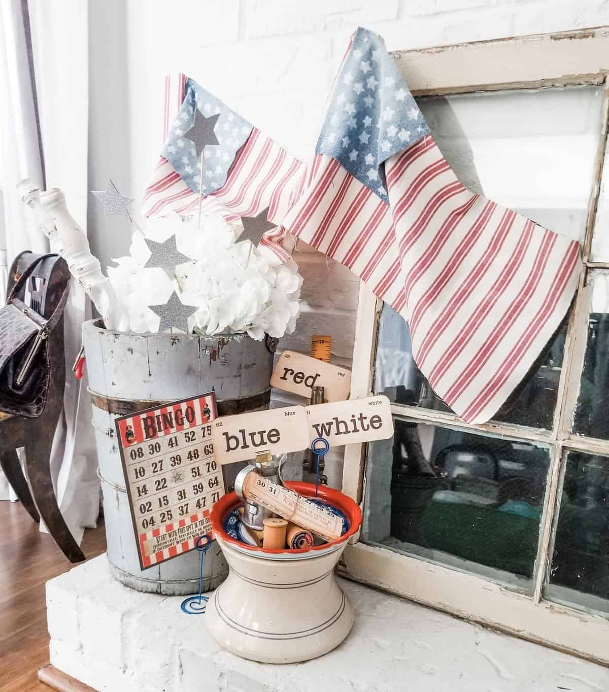 Vintage items and patriotic-themed decor on a rustic mantel, featuring American flag stockings, a bingo card, a pail with flowers and star wands, and a container with spools labeled "red," "blue," and "white.