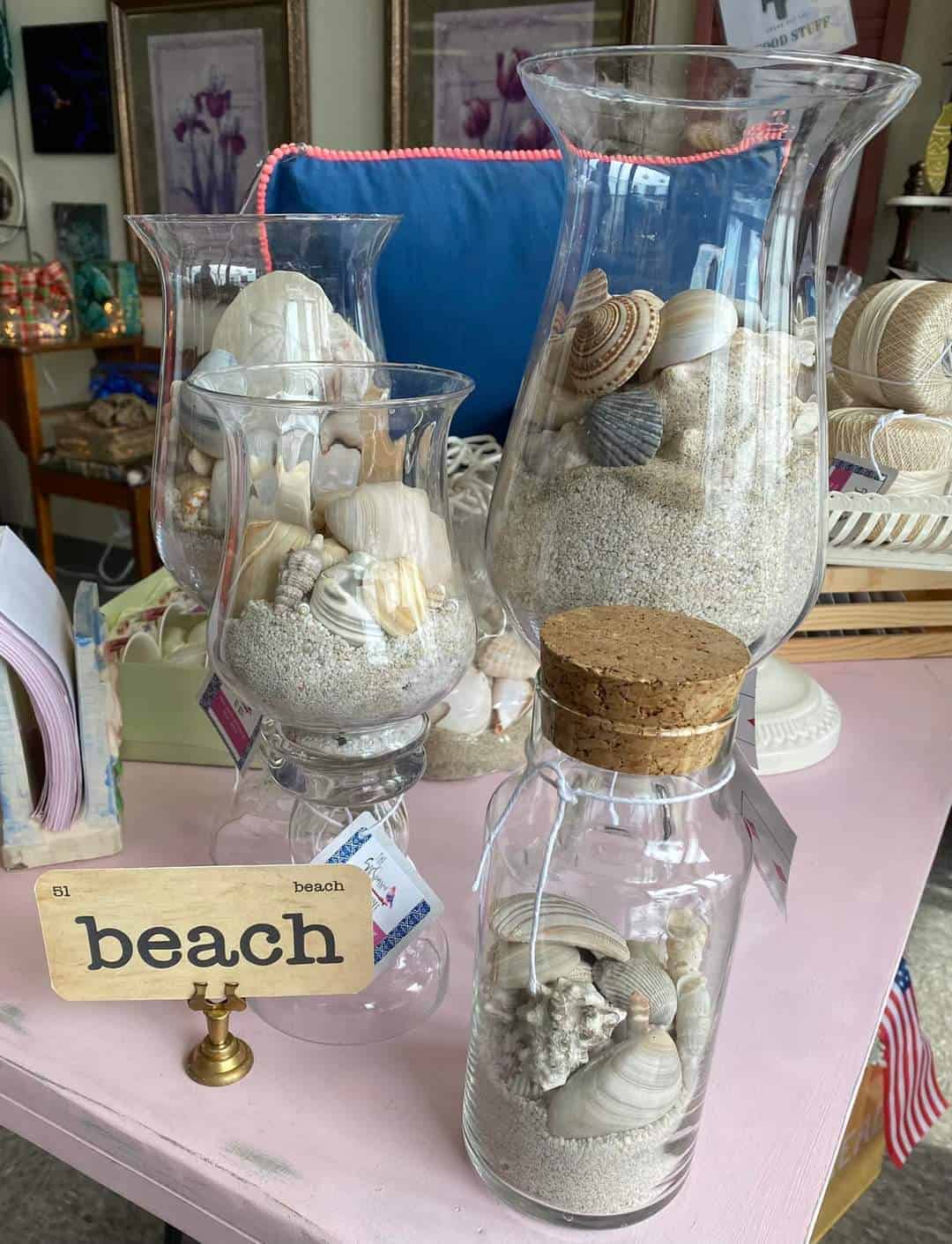 A display of various glass containers filled with sand, seashells, and decorative items labeled "beach," placed on a pink table in a store.