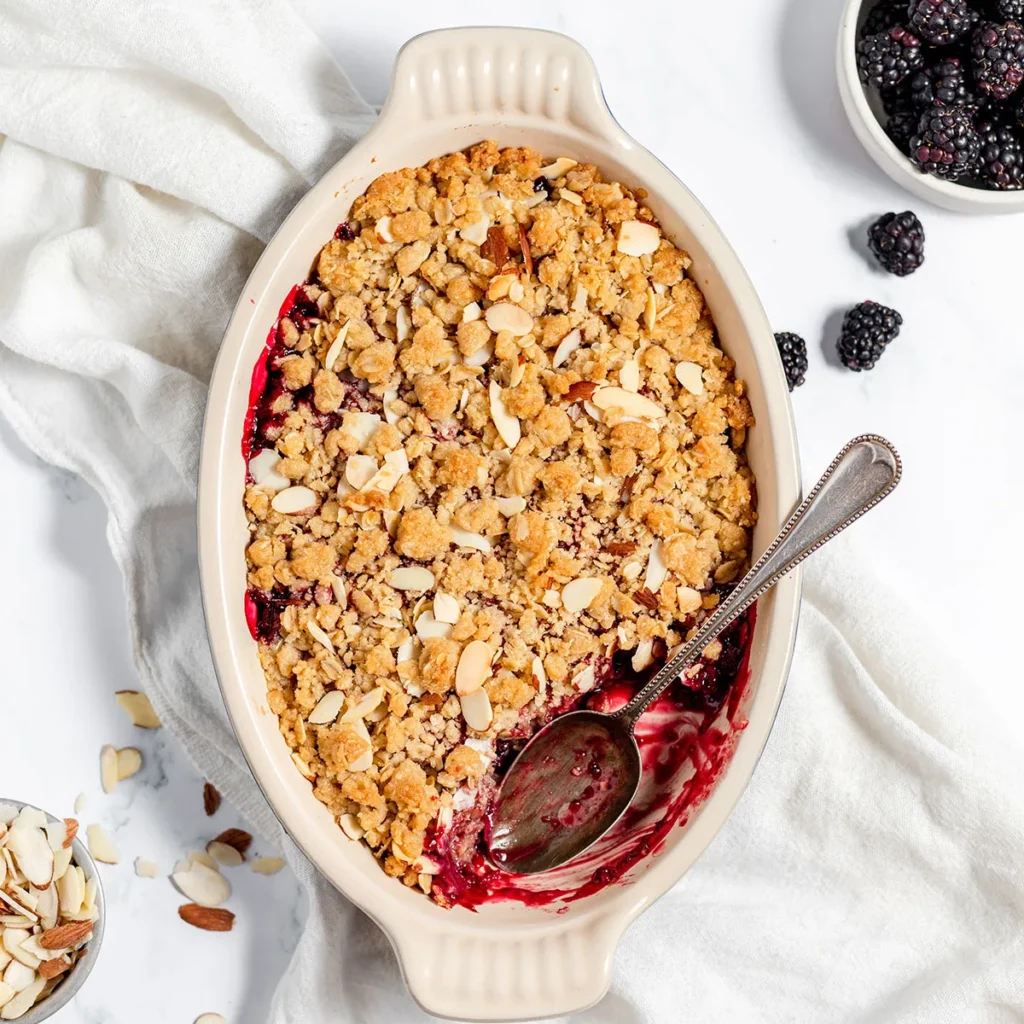 An oval baking dish with a berry crumble topped with a streusel and sliced almonds. A serving spoon is inserted into the dish, revealing the berry filling underneath. Nearby are blackberries and sliced almonds.