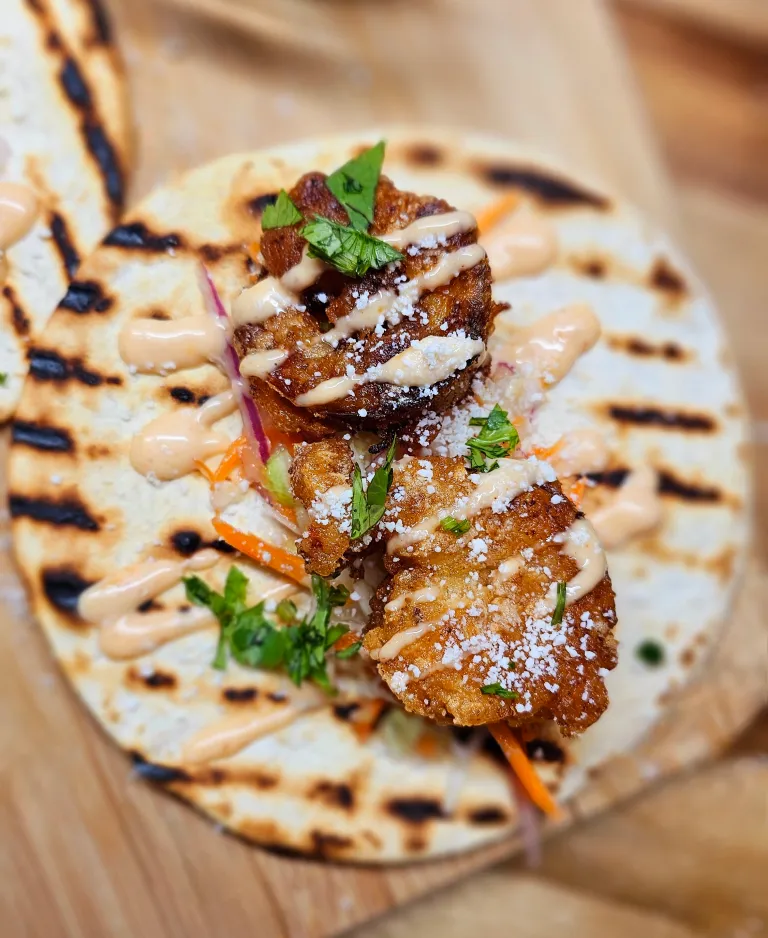 Close-up of a grilled tortilla topped with fried chicken pieces, drizzled with sauce, and garnished with fresh herbs and grated cheese.