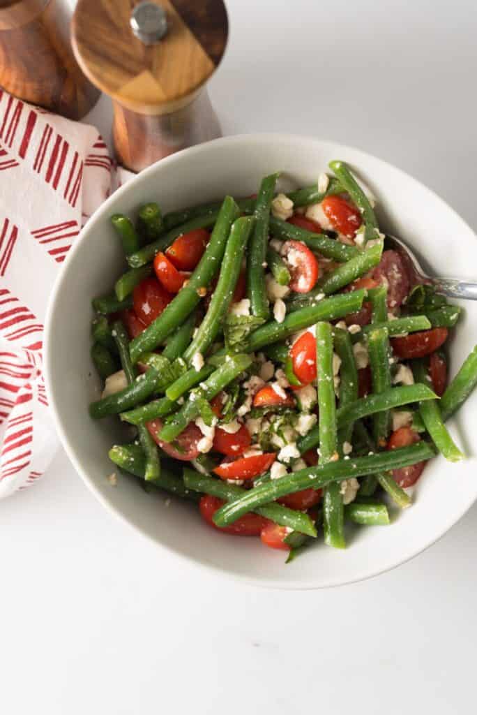 A white bowl contains a salad of green beans, cherry tomatoes, and crumbled feta cheese. A spoon is placed inside the bowl. A red-striped napkin and wooden salt and pepper shakers are near the bowl.