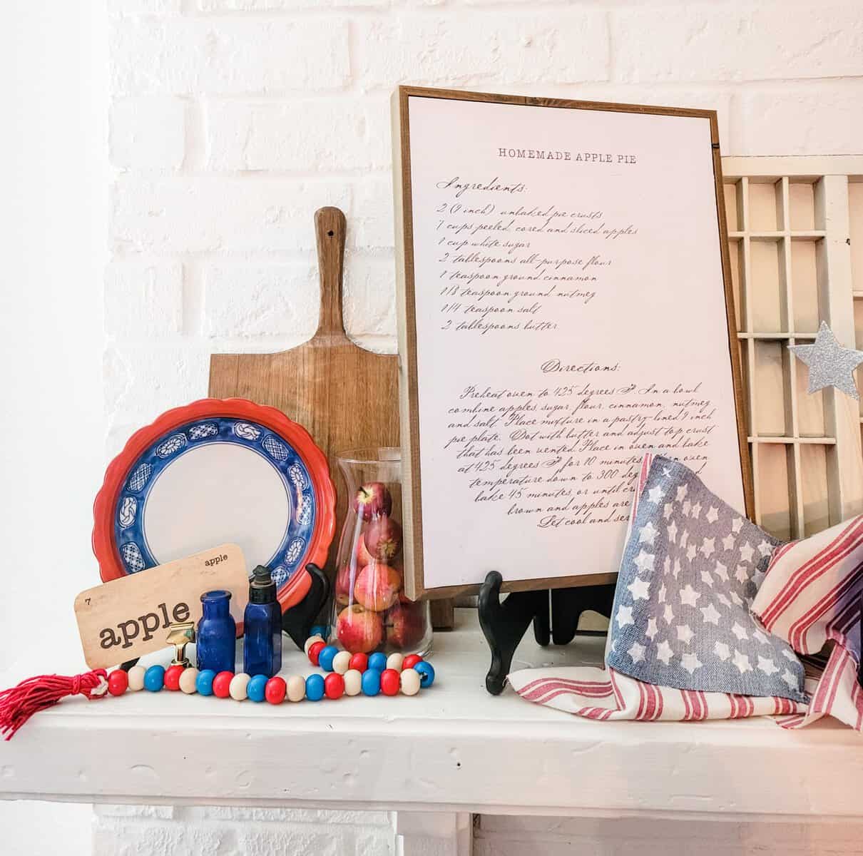 A kitchen shelf displays a homemade apple pie recipe, a cutting board, a decorated plate, apple-themed decor, colorful beads, a fabric star, and a red, white, and blue striped cloth.