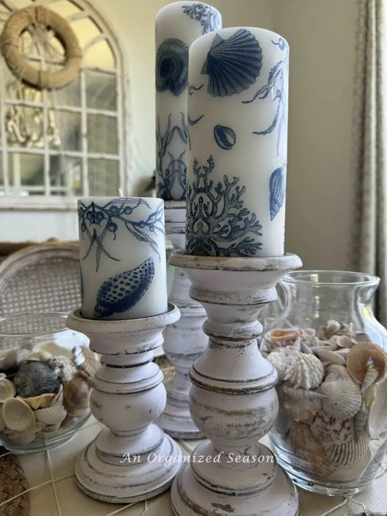 Three white candles with blue seashell and marine life designs on distressed candle holders, surrounded by a variety of seashells in glass containers and a decorative mirror in the background.