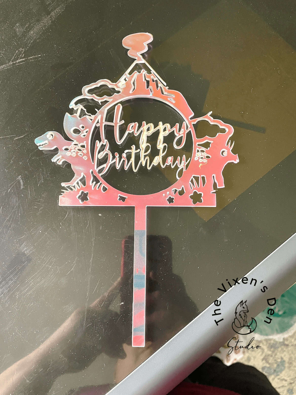 A "Happy Birthday" cake topper with farm animal designs, featuring a cow, pig, and chick, and a barn in the background, placed on a reflective surface.