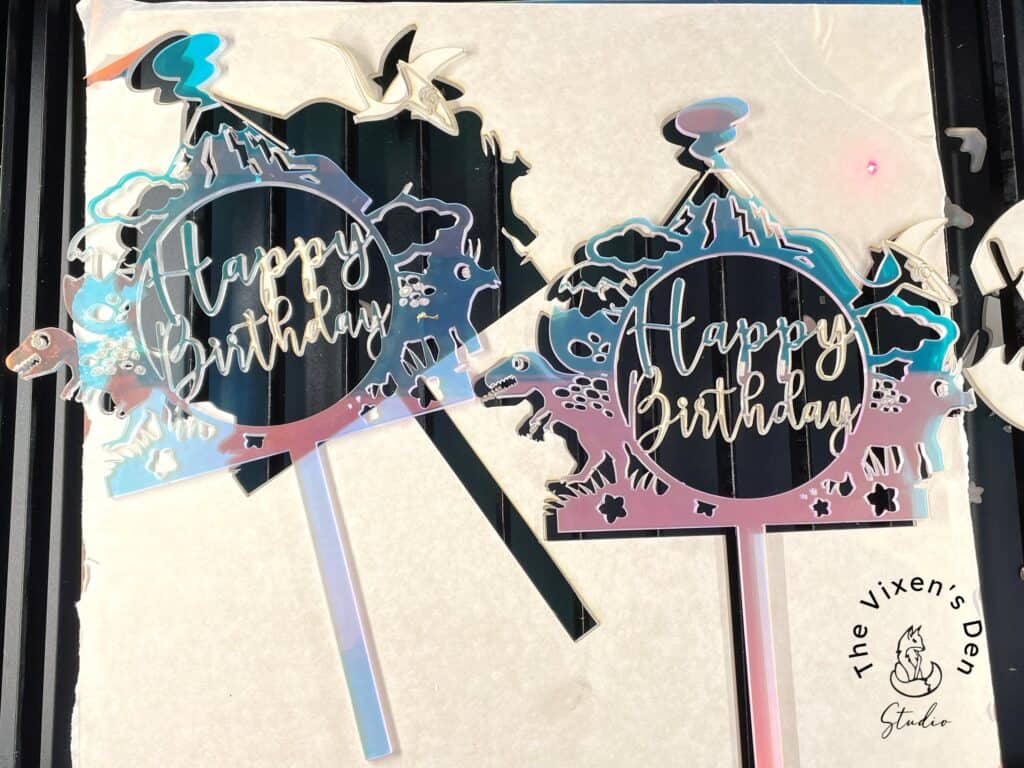 Two iridescent "Happy Birthday" cake toppers with mountain and star designs are placed on a surface. The text "The Vixen's Den Studio" is in the bottom right corner.