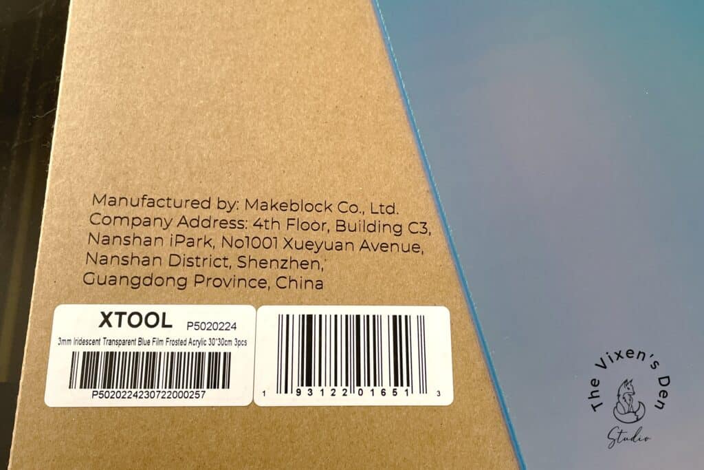 Close-up of a cardboard packaging with manufacturer information for Makeblock Co., Ltd., and product details for a 3mm iridescent blue film. A barcode and a logo reading "The Vixen's Den Studio" are visible.
