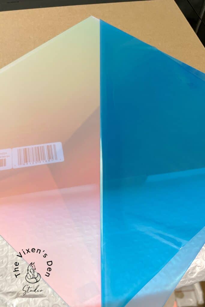 Two sheets of translucent, colored plastic on a table. One sheet is gradient pink to blue with a barcode sticker, and the other is solid blue. Branding "The Vixen's Den Studio" is visible in the corner.
