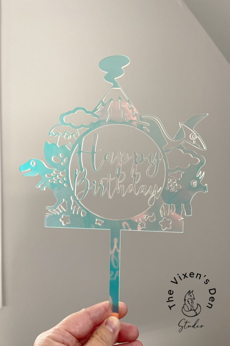 A hand is holding a turquoise cake topper with "Happy Birthday" text, featuring dinosaur designs and the Vixen's Den Studio logo in the bottom right corner.