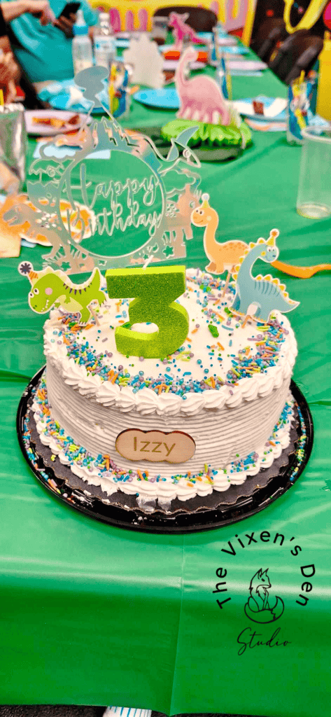 A birthday cake topped with colorful dinosaur decorations and a green "3" sits on a table with party items and people in the background. The cake has the name "Izzy" on an oval plaque.