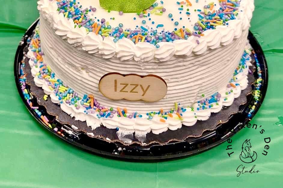 A round cake with white frosting, colorful sprinkles, and a green decoration on top. A label on the front reads "Izzy." The cake is on a black tray placed on a green tablecloth.