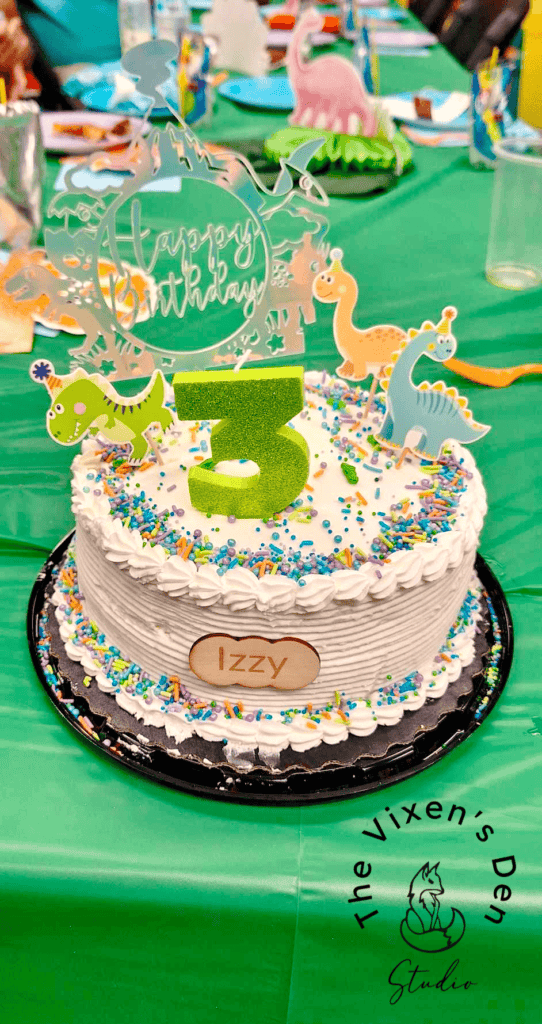 A white-frosted birthday cake with colorful sprinkles, dinosaur decorations, a "Happy Birthday" topper, and a large green number "3." A name plaque reads "Izzy." The cake is on a green tablecloth.
