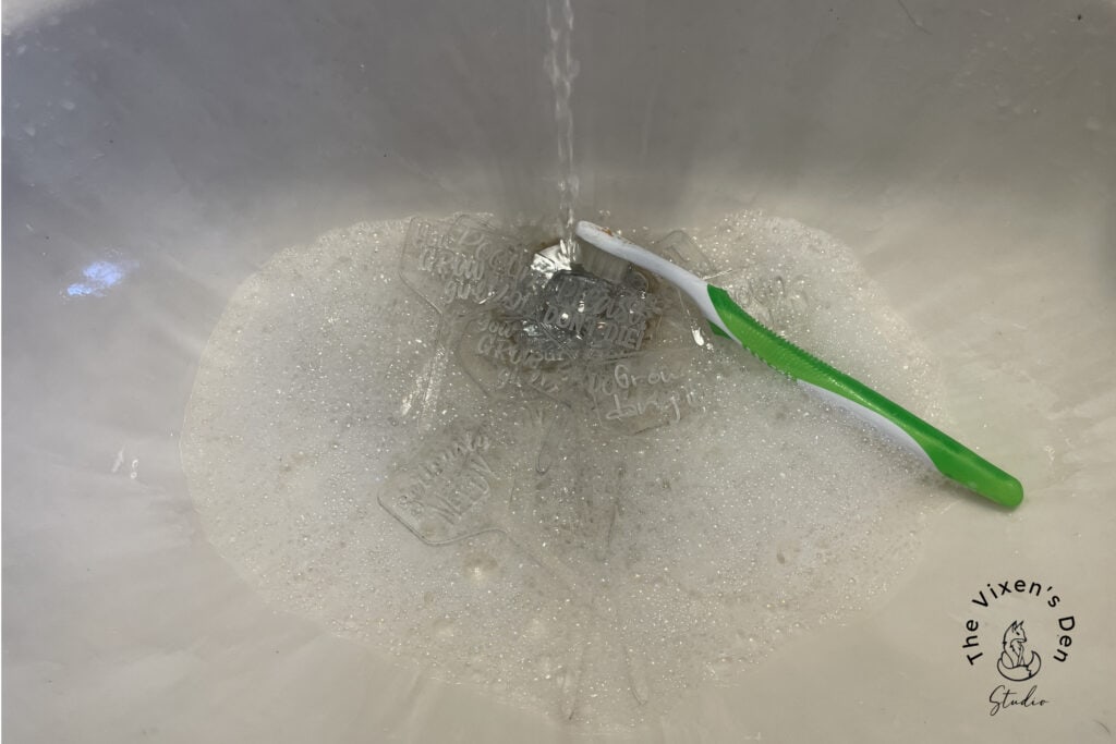 A green toothbrush lies in a soapy sink under running water, alongside a few small translucent objects with text that are being cleaned. The Vixen's Den Guide logo is visible in the bottom right corner.