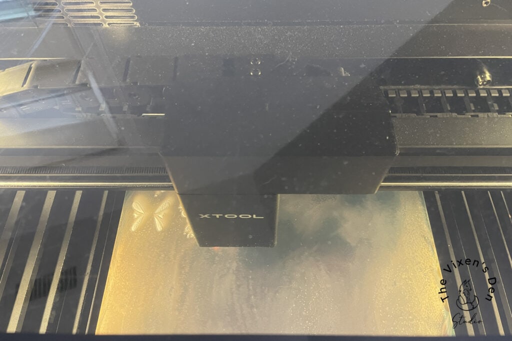 A top-down view of a laser engraver machine in operation, with the label "XTOOL" visible on the device. The machine is engraving a shiny, flat surface. A logo is present at the bottom right corner.