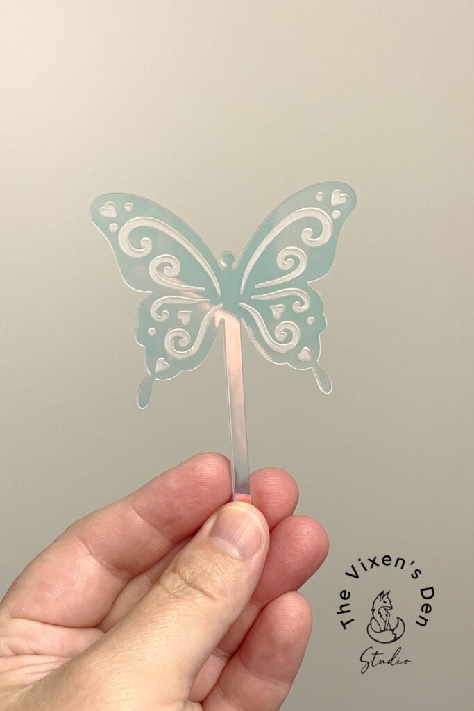 A hand holds a clear, decorative butterfly-shaped object with intricate cutouts. Below, a logo reads "The Vixen's Den Studio.