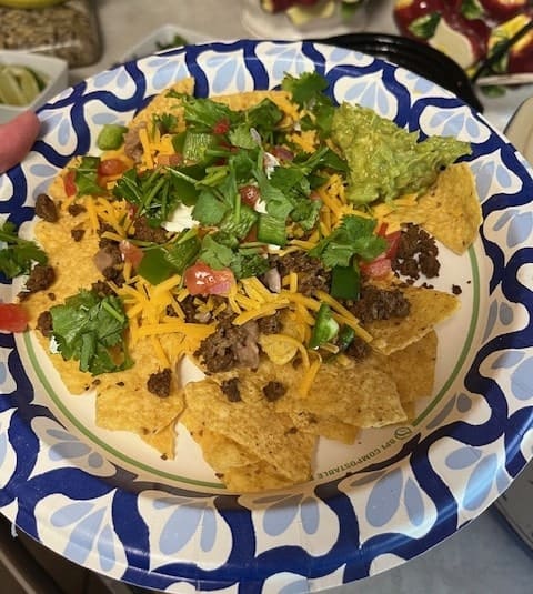 A paper plate with nachos topped with ground meat, shredded cheese, diced tomatoes, jalapeños, cilantro, and a side of guacamole.