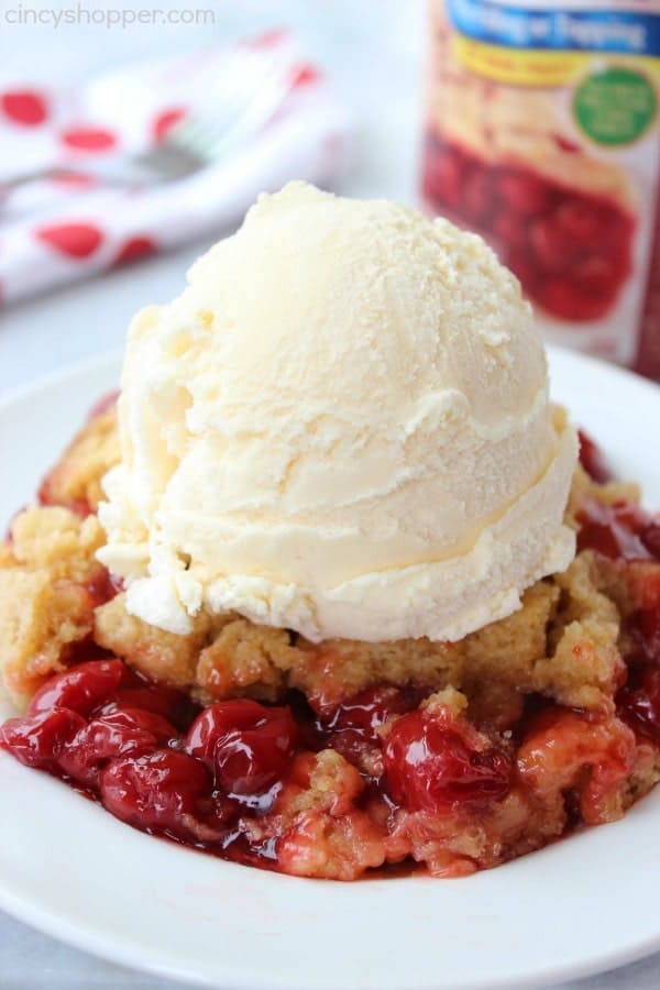 A plate of cherry cobbler topped with a scoop of vanilla ice cream, with a container of cherry pie filling in the background.