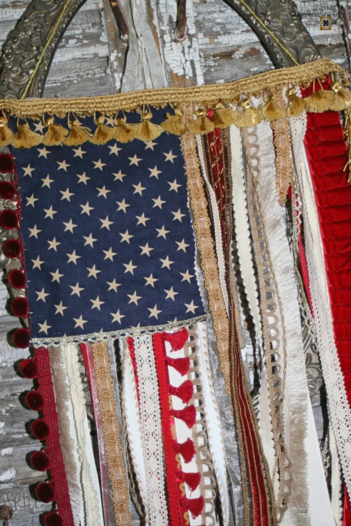 A fabric decoration features an American flag pattern with numerous strips of various materials, including lace and ribbon, hanging from it.