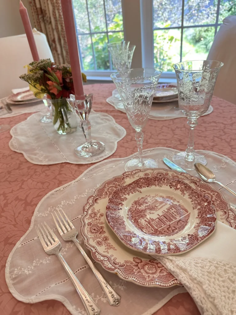 A dining table set with pink candles, crystal glasses, floral arrangements, and vintage red-and-white patterned plates on a pink tablecloth. Natural light comes in through a nearby window.