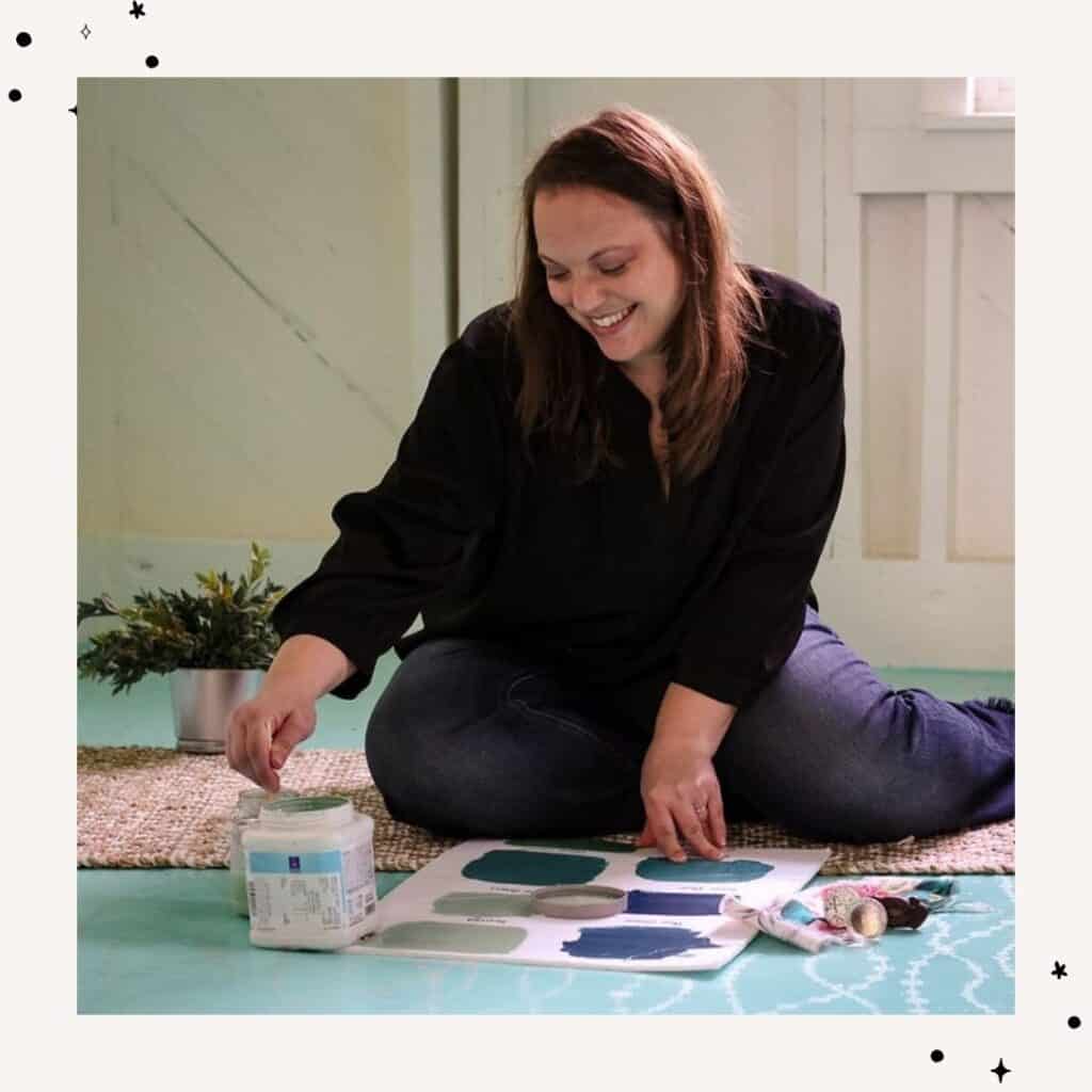 A woman sits on the floor, smiling, as she works with paint samples and a paint container on a piece of paper. She is surrounded by a plant and some additional artistic supplies.