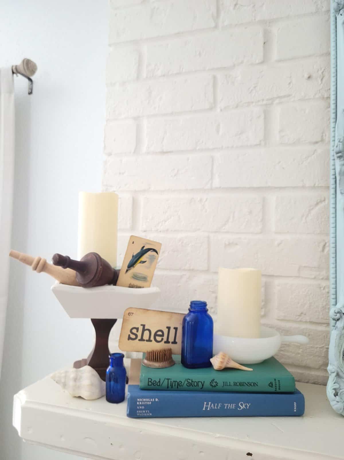 A white shelf displays candles, books, blue bottles, a shell, a honey dipper, and a word card spelling "shell," with a white brick wall background.