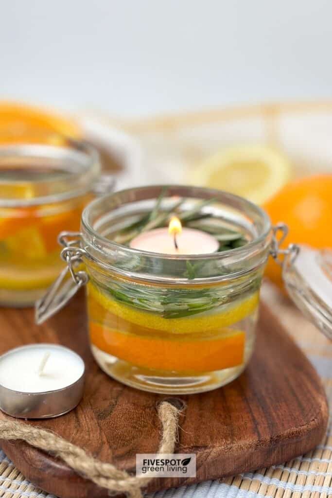 Two small jars filled with citrus slices and herbs, each topped with a lit tea light candle, placed on a wooden board. A lemon and another jar are in the background.