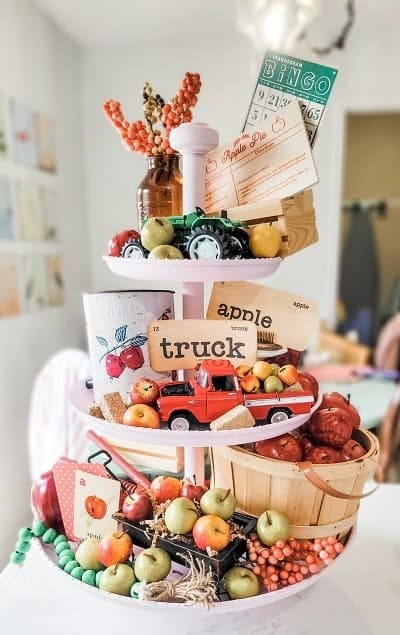 A three-tiered wooden stand displaying apples, a toy tractor and truck, bingo cards, a basket of small pears, and various autumn-themed decorations.