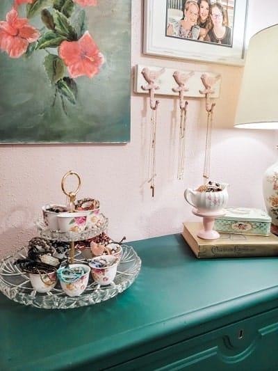 A teal dresser with a multi-tiered dish holding jewelry, a lamp, and books. Above, there is a framed photo and a decorative floral hook rack with necklaces hanging from bird-shaped hooks.