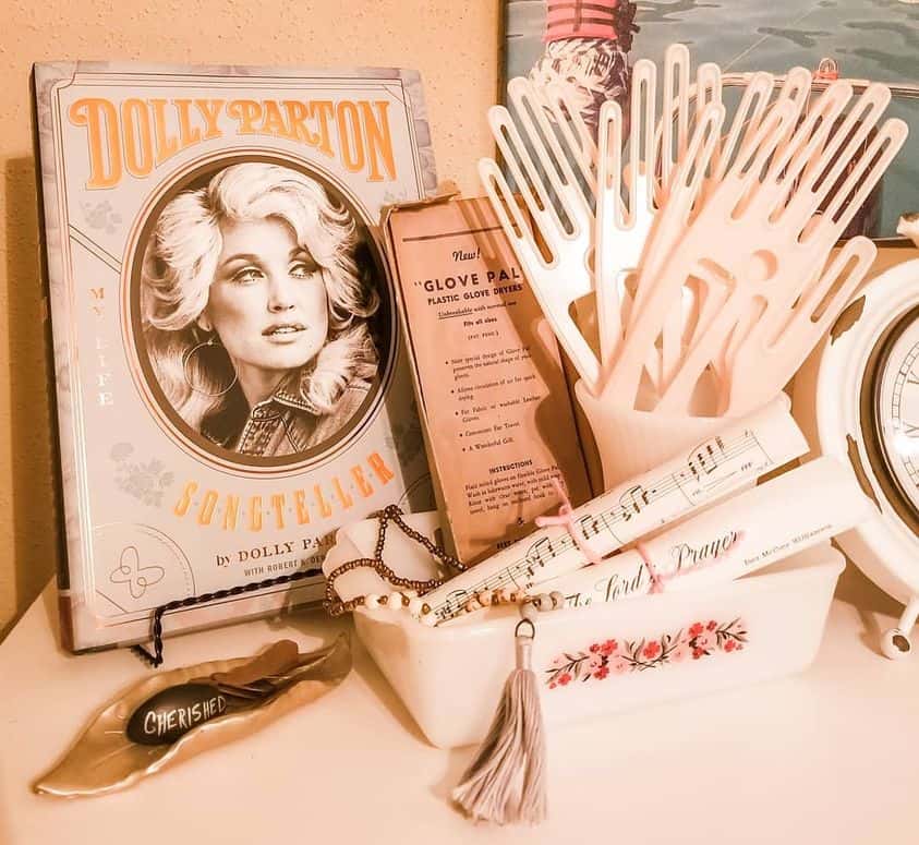 A vanity display featuring a Dolly Parton book, a vintage "Glove Palm" box, plastic combs, a "Cherished" leaf trinket dish, a musical scroll, and a decorative tassel in a small floral box.