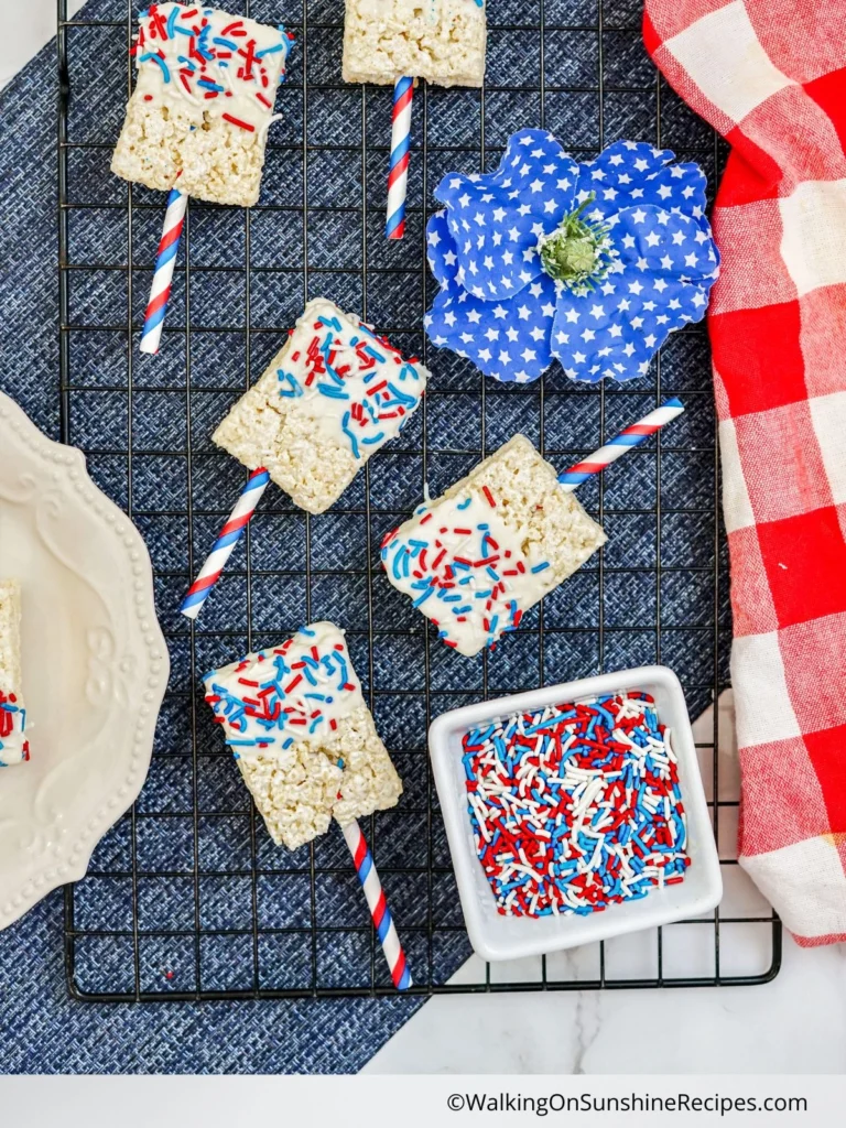 Rice Krispies treats on sticks with red, white, and blue sprinkles, placed on a cooling rack, next to a bowl of sprinkles, a blue flower, and a red-and-white checked cloth.