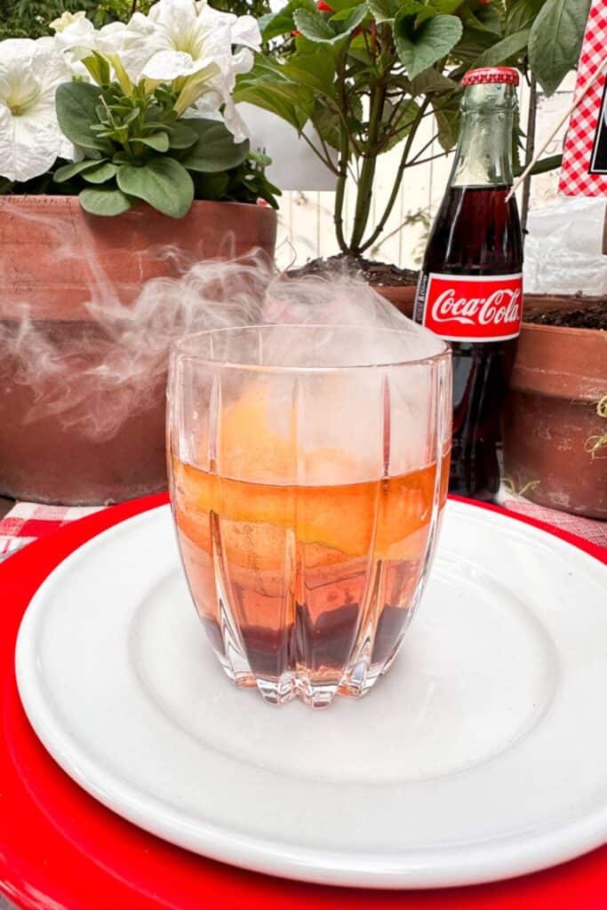 A glass with a smoking cocktail on a white plate with red trim, accompanied by a bottle of Coca-Cola and potted plants in the background.