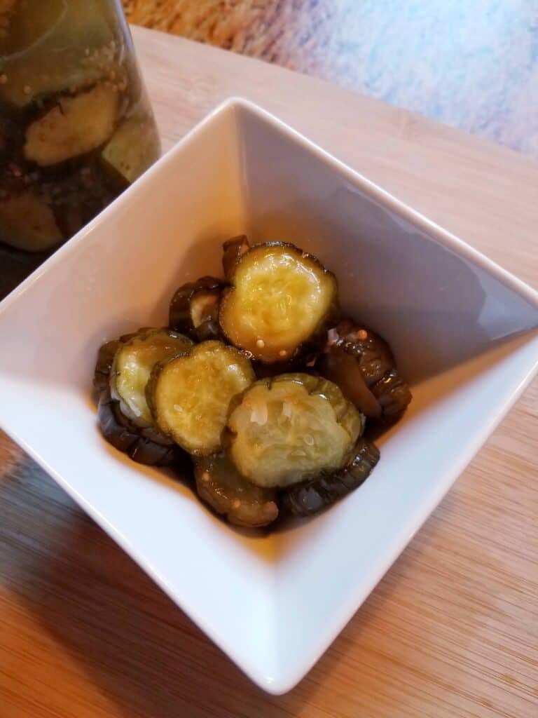 A white square bowl containing sliced pickles, placed on a wooden surface.