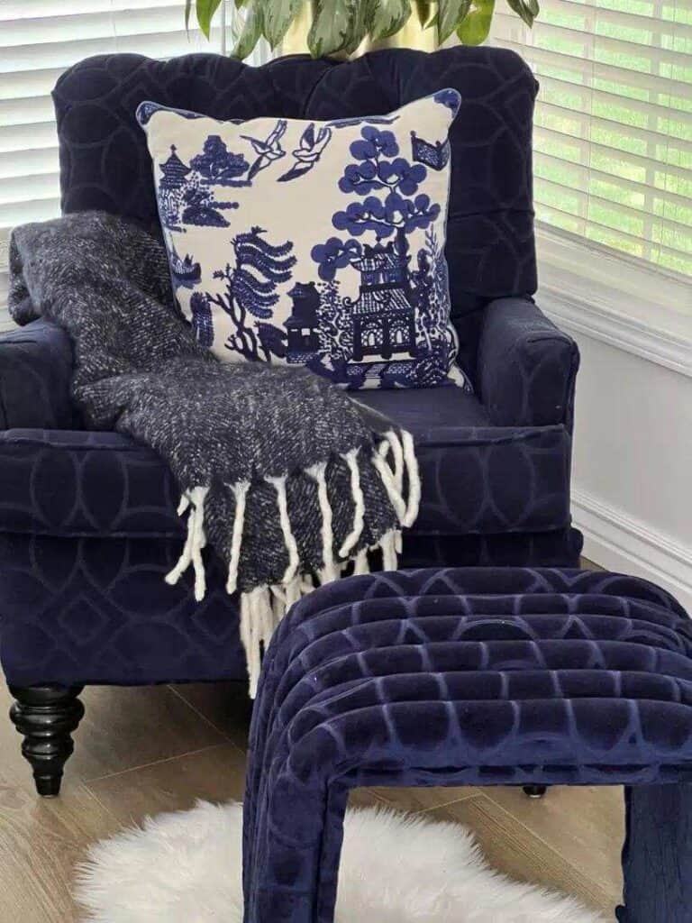 A navy blue armchair with a matching ottoman, draped with a gray throw blanket and a white pillow featuring blue patterns, placed in a room with white blinds and wooden flooring.