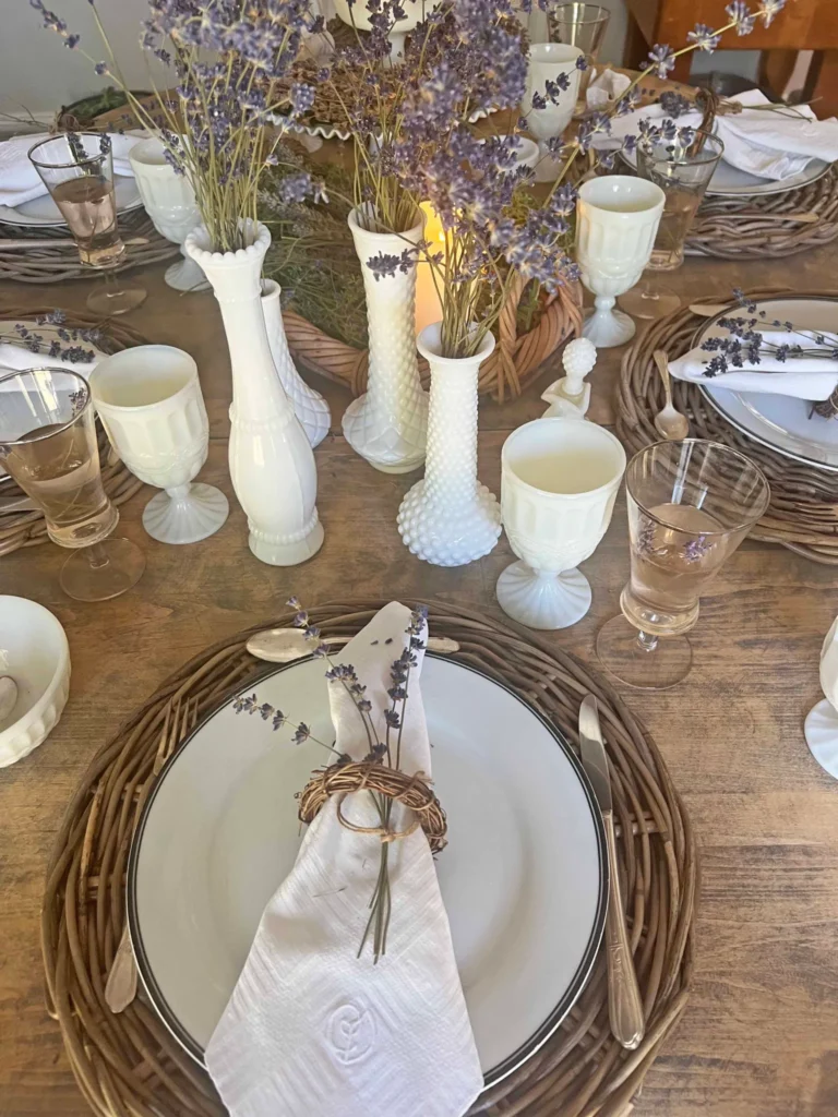 A rustic dining table set with white plates, woven chargers, and lavender sprigs in glassware. Milk glass vases filled with lavender serve as centerpieces. Neutral-toned glasses and napkins are also present.