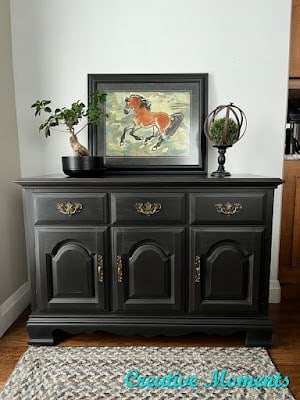 A black wooden sideboard with brass handles has a framed horse painting, a small leafy plant, and a round decorative item on top. A patterned rug lies in front. The text "Creative Moments" is visible in the image.