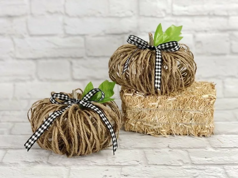 Two decorative pumpkins made of twine with green leaves and black and white checkered ribbon accents are placed on a hay bale against a white brick background.