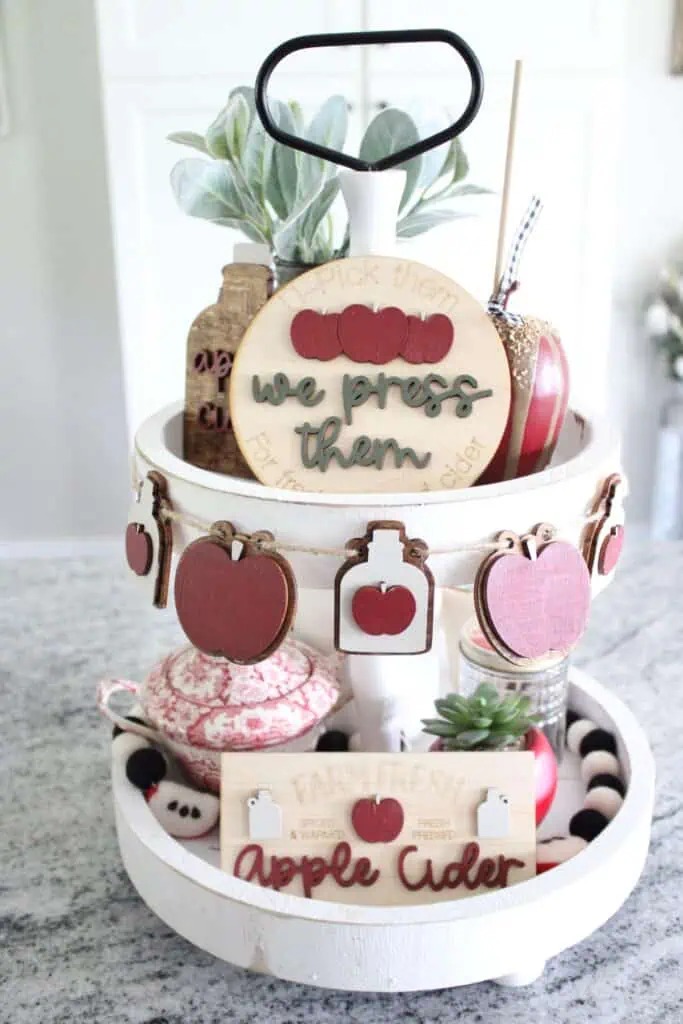 A two-tiered tray with autumn-themed decorations, including wooden apple signs, a potted plant, apple cider labels, and a ceramic dish, set on a kitchen counter.