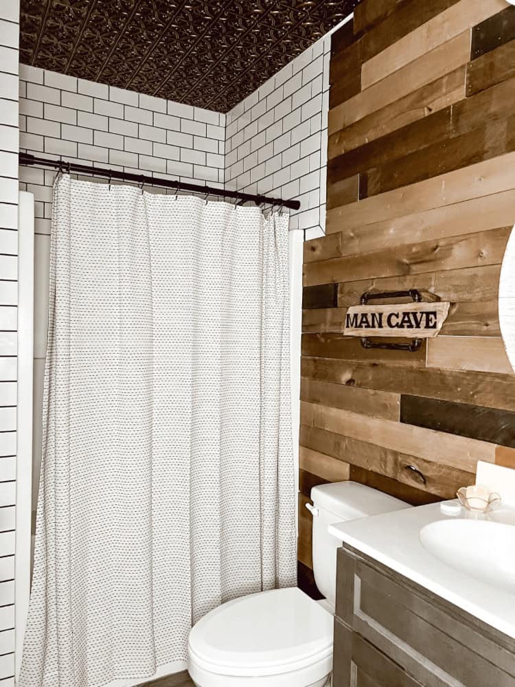 A bathroom with white tiled walls, wooden panel accent wall, a shower with a curtain, a toilet, and a vanity sink. A sign reading "Man Cave" is mounted on the wooden wall above the toilet.