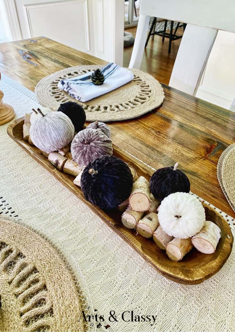 A wooden table is set with woven placemats, cloth napkins, and a rectangular wooden tray holding decorative fabric pumpkins and birch wood pieces.