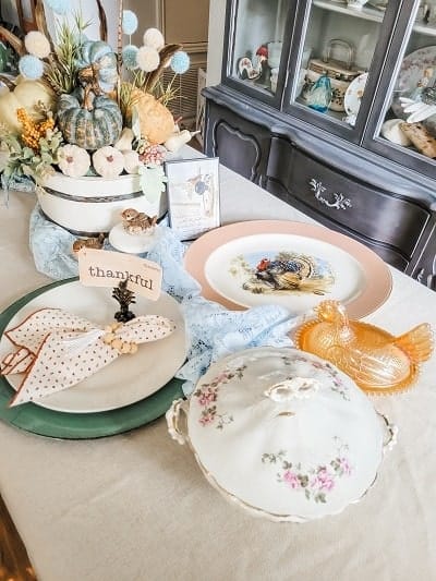 A table is set with autumn-themed decor, including a "thankful" sign, a floral dish, a turkey platter, and a chicken-shaped glass dish. A decorative bouquet and a china cabinet are in the background.