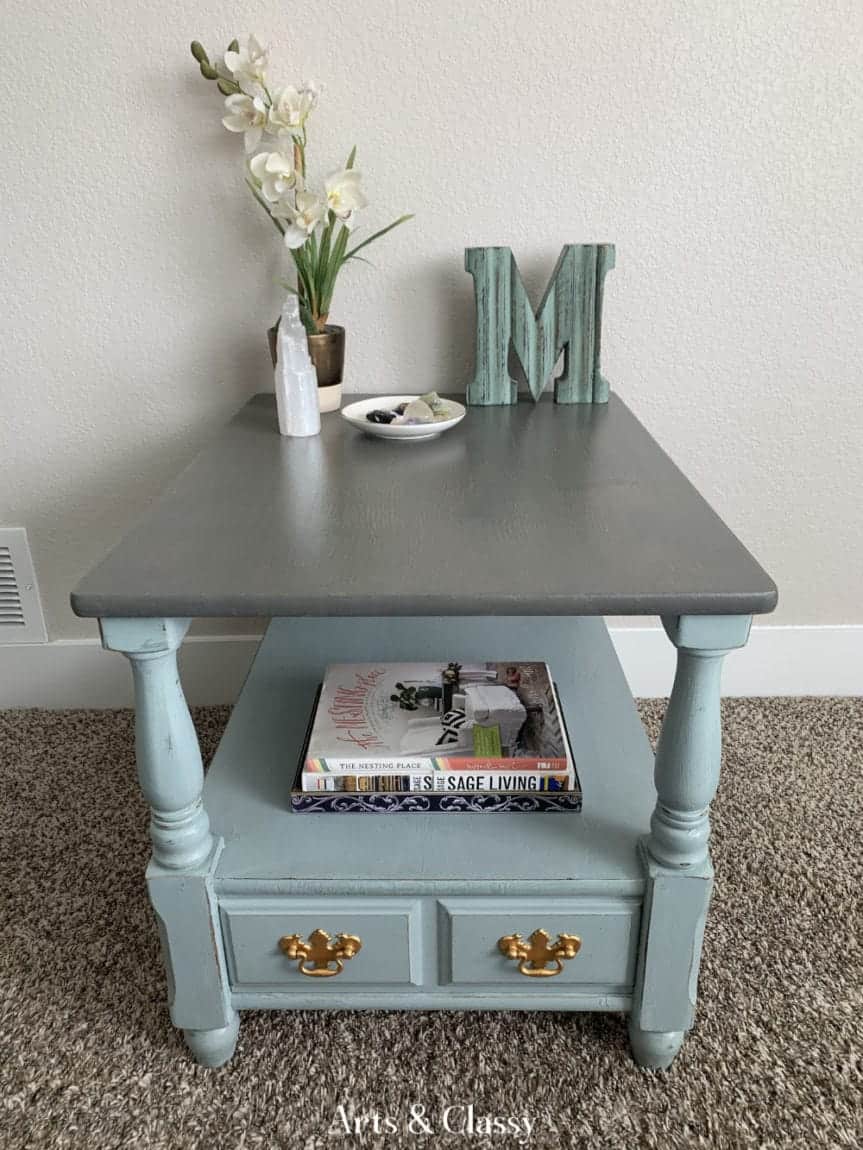 A small side table with a gray top and light blue base, featuring a drawer with gold handles. The top holds an orchid, a decorative letter "M", a small dish, and books are on the lower shelf.