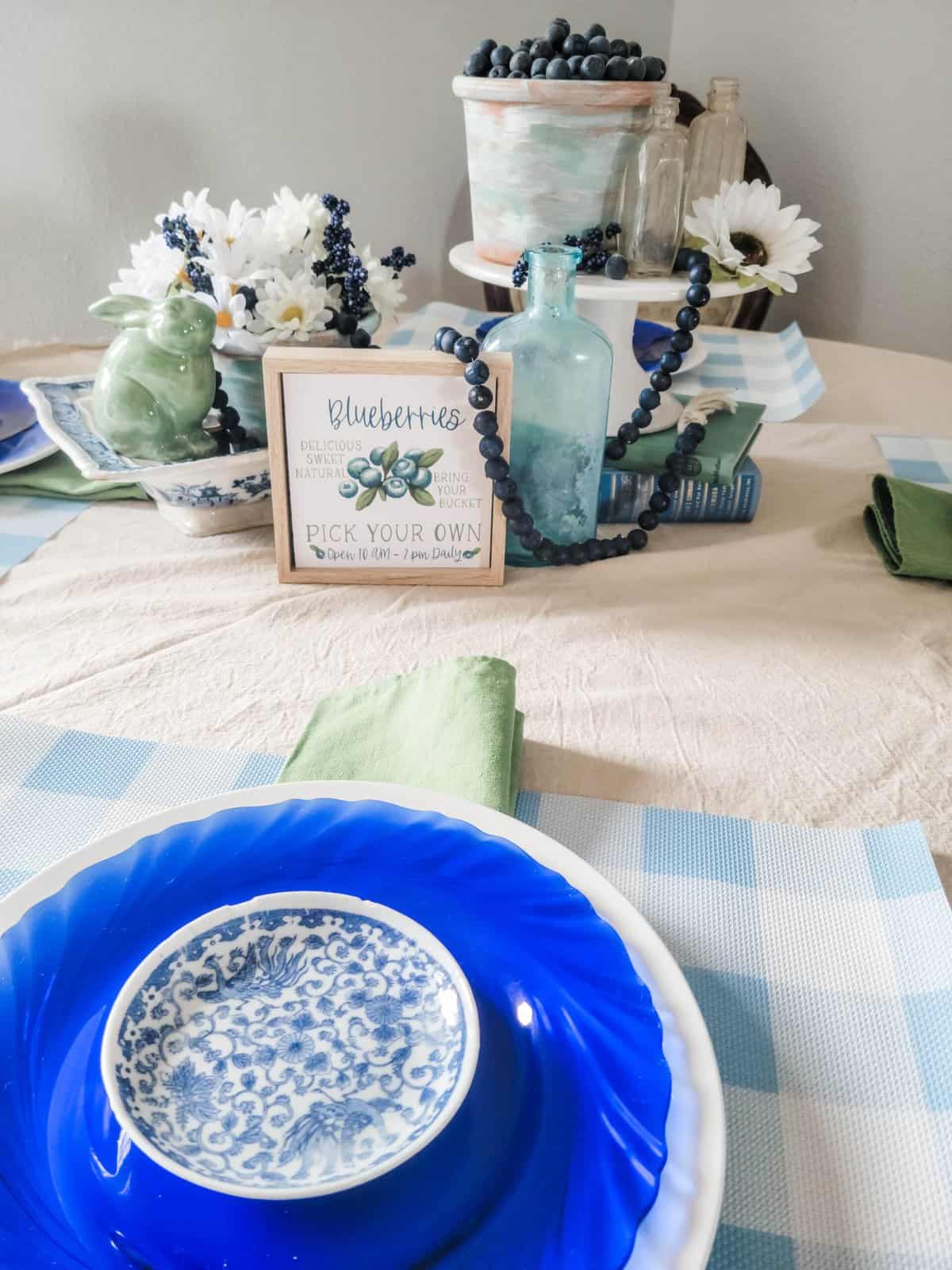 A table set with blue and white dishes, green napkins, and blueberry-themed decorations, including a framed blueberry sign, ceramic bird, and a potted plant with blueberries.