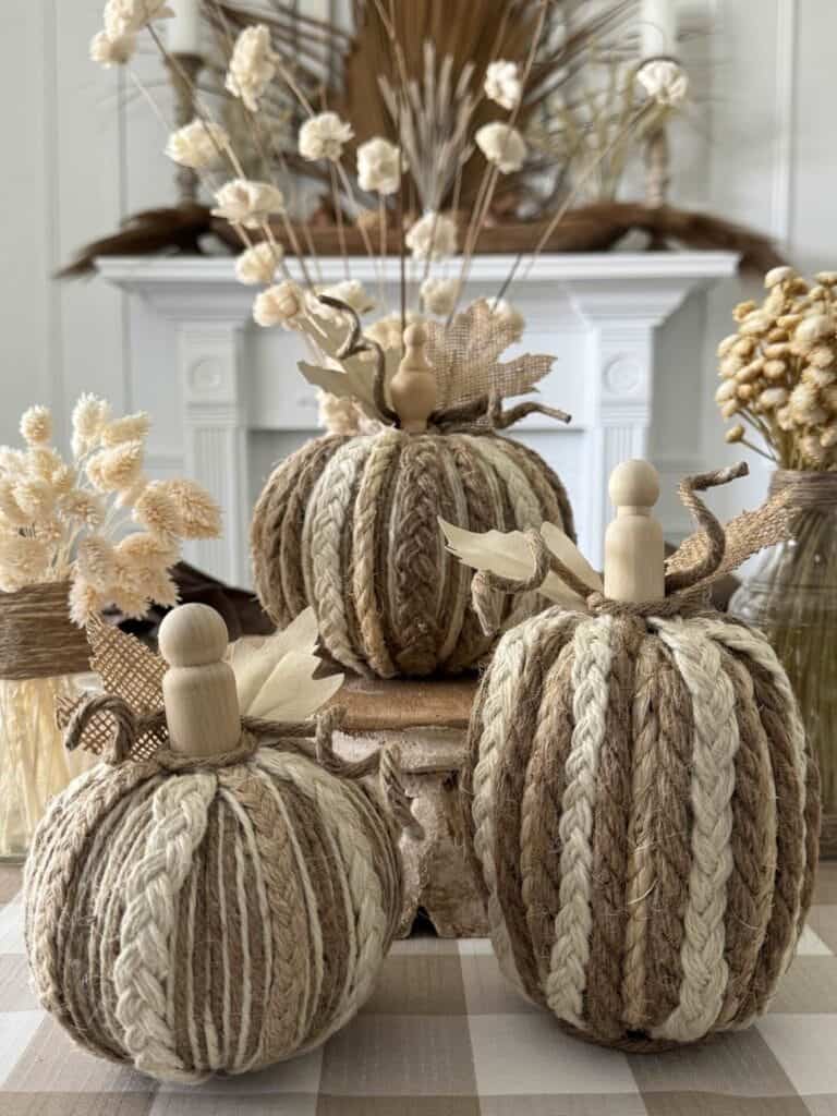 Three decorative pumpkins made from braided twine, each topped with a small wooden stem, are displayed on a checkered cloth in front of a fireplace adorned with dried floral arrangements.