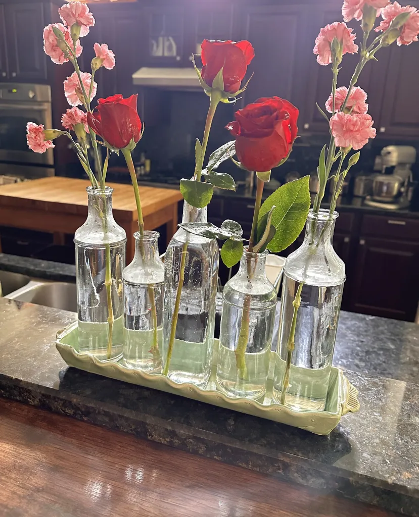 Clear glass bottles arranged in a ceramic tray hold a mix of pink carnations and red roses, placed on a dark kitchen counter with a blurred background of wooden cabinets and kitchen appliances.