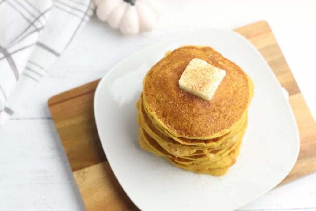 A stack of pancakes topped with a pat of butter, placed on a white plate on a wooden cutting board. A white pumpkin and a white checked napkin are in the background.