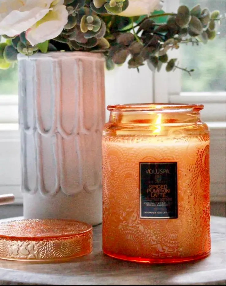 An orange-tinted candle jar labeled "VOLUSPA Spiced Pumpkin Latte" is lit and placed on a table beside a decorative vase with flowers.