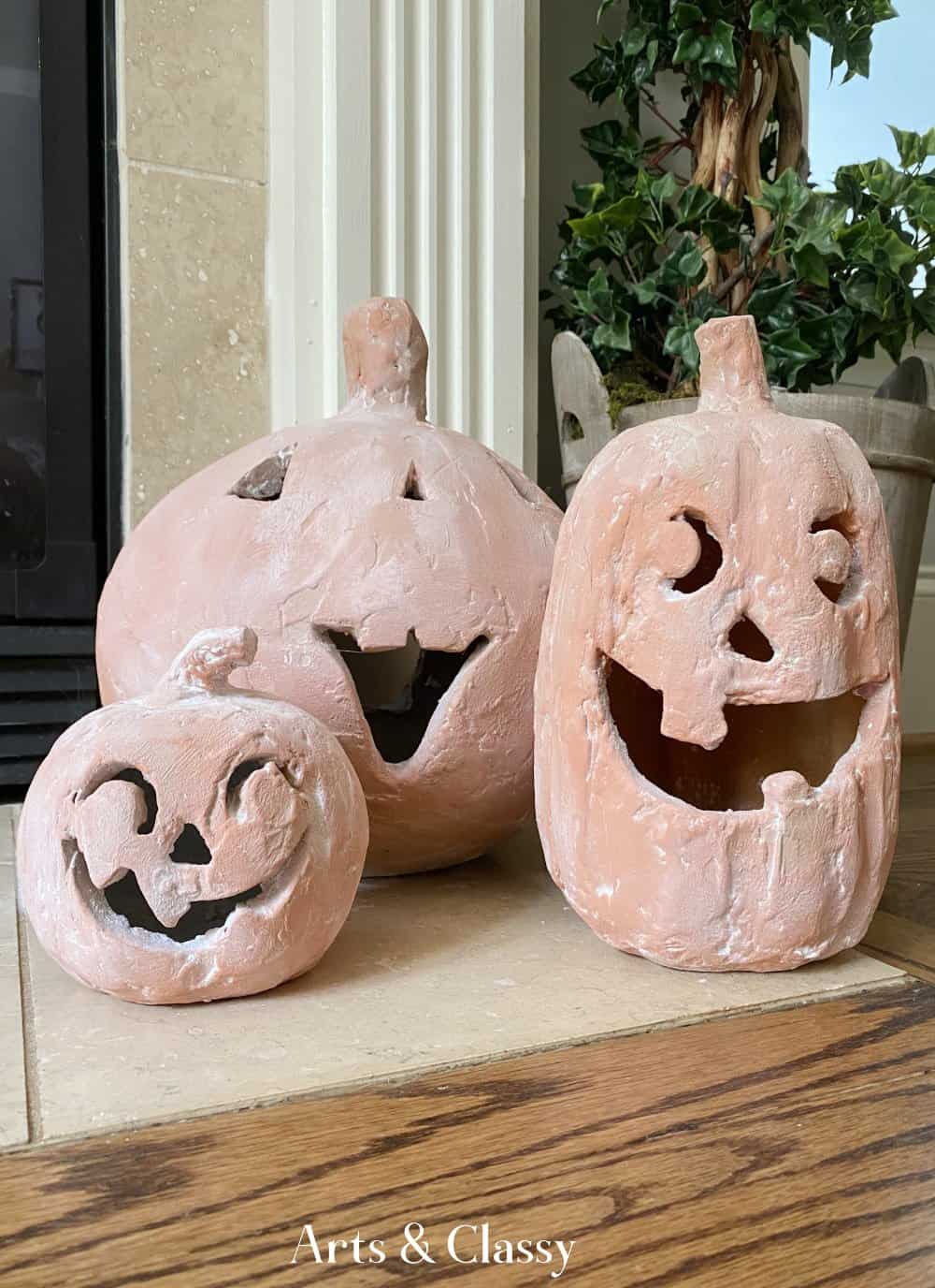 Three clay pumpkin lanterns with carved faces sit on a wooden floor next to a fireplace and potted plant. The pumpkins have various expressions and appear to be for Halloween.