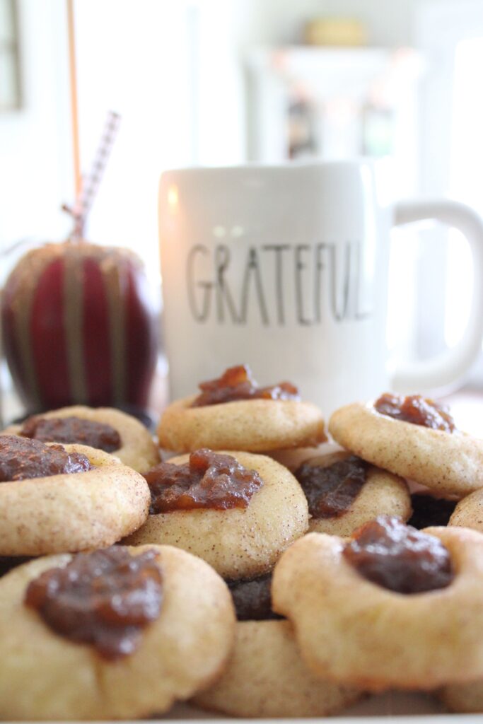 A pile of cookies topped with apple jam arranged near a white mug with "GRATEFUL" written on it. In the background, there is an apple-shaped decoration with a black straw.