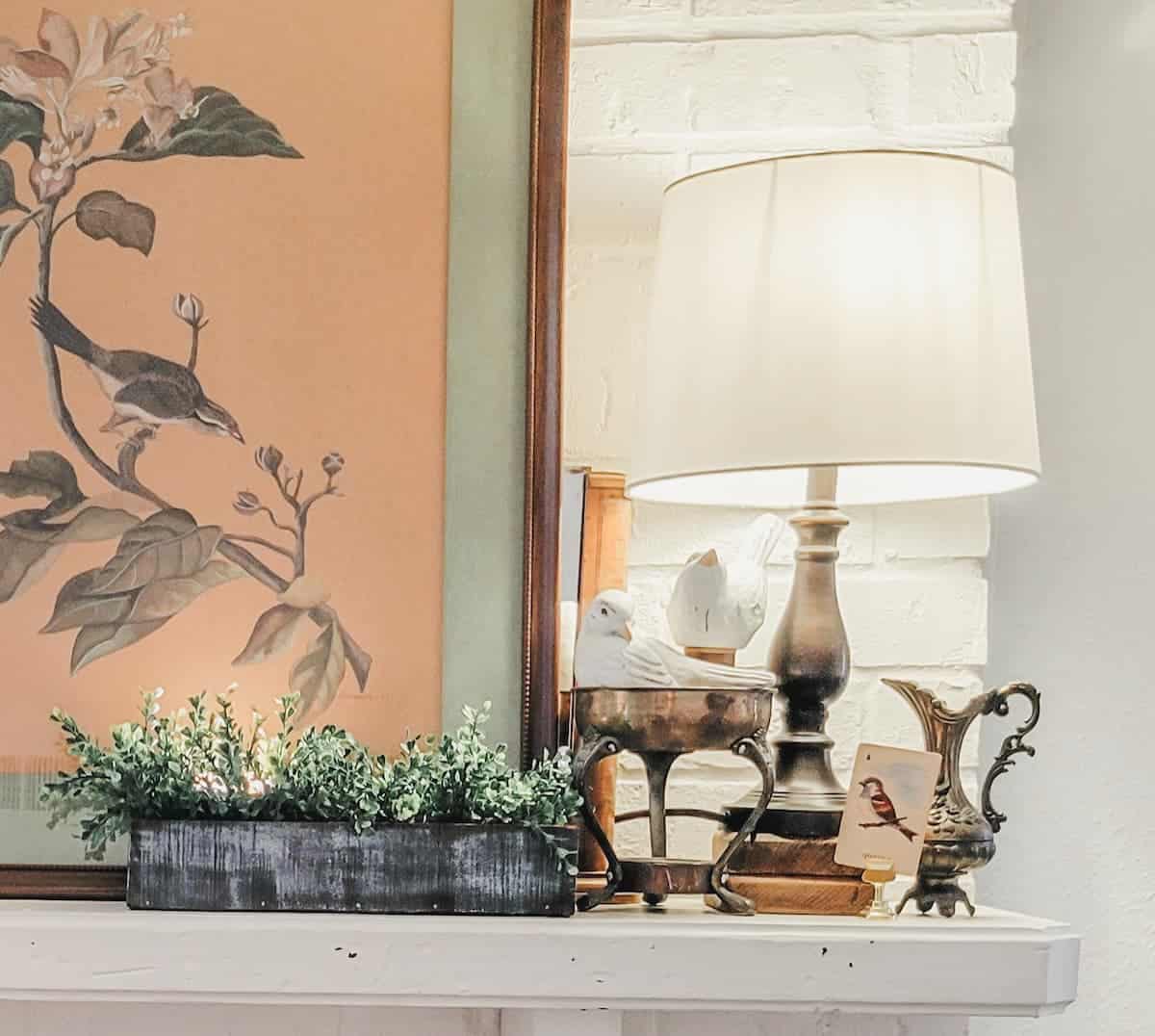 A shelf with potted greenery, a framed bird artwork, a lamp, decorative figurines, and a small ornamental container on a white brick background.