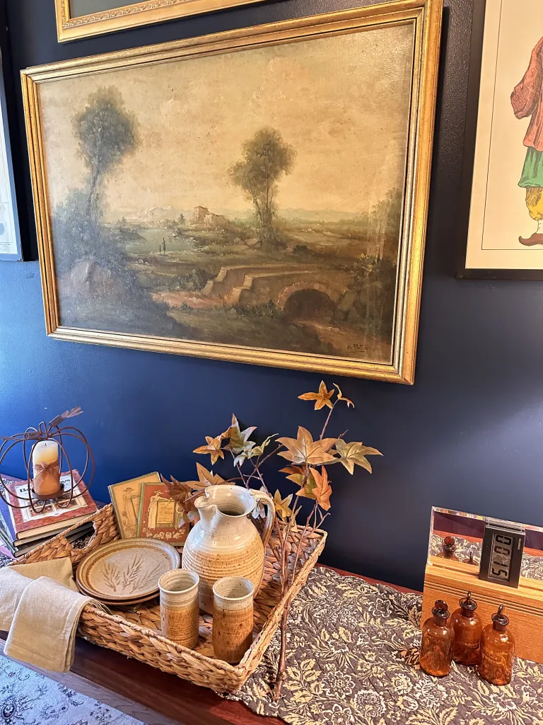 A rustic display featuring a large landscape painting, wicker tray holding pottery items, dried flowers, old books, and three amber bottles against a dark wall.