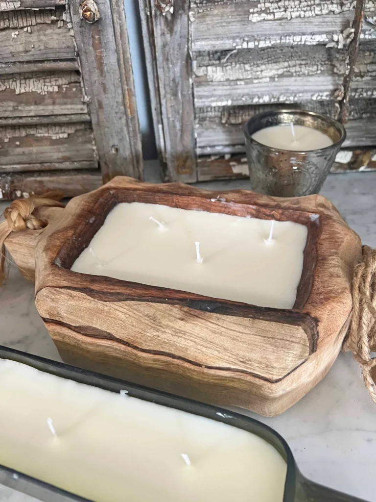 A wooden candle holder with four white candles, a small candle in a metal holder, and another candle in a black rectangular holder placed on a rustic wooden surface.